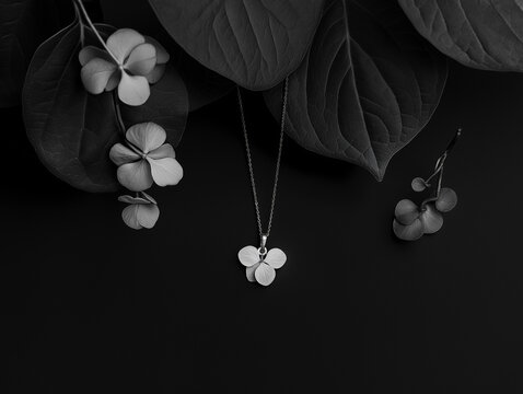 A silver necklace with a flower pendant sits on a black background. The necklace is surrounded by leaves, which add a natural touch to the scene. Scene is elegant and serene, with the necklace
