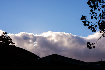 A beautiful blue sky with light gray and white clouds forms a backdrop for silhouetted black rooftops, set against backlighting, with tree branches adding depth to the tranquil rural scene