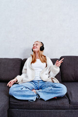 A woman relaxes on the sofa, enjoying music through her headphones. An attractive young girl takes advantage of her leisure time for rest and enjoyable activities at home.