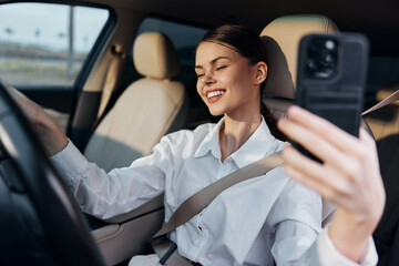 A woman sits in the driver's seat of a car, taking a selfie with her cell phone, holding it up to the camera with a focus on the selfportrait concept