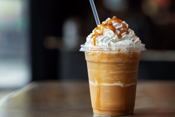 caramel frappuccino smoothie in a clear plastic cup with a straw, finished with a swirl of whipped cream and a drizzle of caramel sauce, set on a cafe table