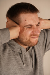 Portrait of young guy closing his ears by hands and keeping his eyes closed while isolated on grey. Photo of desperate annoyed bearded man keeps hands on head and closes eyes. Man in depression.`