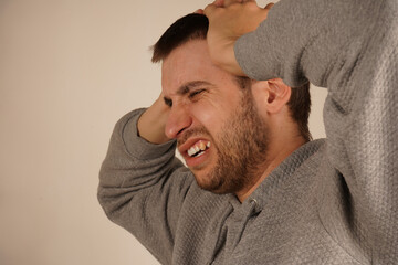 Fototapeta premium Man in depression. Portrait of young guy closing his ears by hands and keeping his eyes closed while isolated on grey. Photo of desperate annoyed bearded man keeps hands on head. Man with headache.