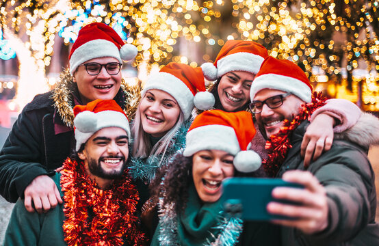 Fototapeta Self portrait of young people wearing santa claus hat celebrating Christmas day outside - Happy multiracial friends group taking selfie under tree having xmas holiday party - Winter holidays concept