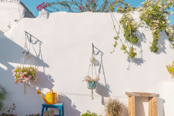 In a picturesque courtyard on Paros, vibrant hanging flower pots and a bright yellow watering can complement the whitewashed wall, creating a serene atmosphere beneath the sunny sky.