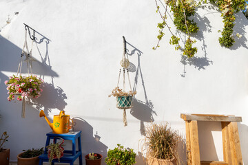 A sunlit wall in Paros features two hanging planters filled with flowers, accompanied by a yellow watering can, creating a welcoming outdoor atmosphere.