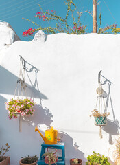 Hanging flower pots filled with blooms complement a whitewashed wall on Paros. A cheerful yellow watering can and wooden furniture enhance the garden ambiance in the Cyclades.
