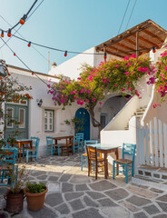 A picturesque alley in Paros features a traditional house surrounded by bright bougainvillea. Outdoor tables create a welcoming atmosphere for visitors to relax and enjoy the view.