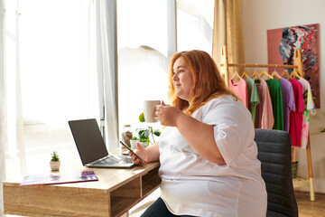 A vibrant plus size woman sips coffee while working on her laptop, surrounded by colorful clothes.