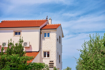 Modern Mediterranean house with red tile roof, white stone facade, and lush greenery under bright blue sky. Residential building at summer day