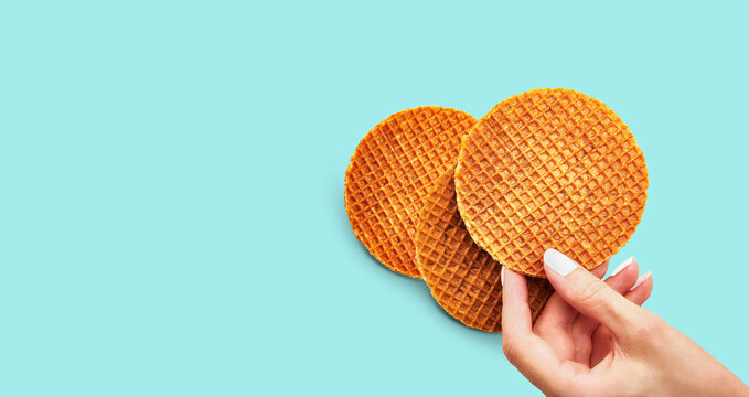 Female's hand holding stroopwafel over blue background. Traditional Dutch waffle caramel cookie. Delicious snack concept, closeup shot, flat lay, horizontal orientation, selective focus, long banner