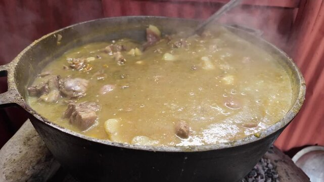Close-up of a steaming pot cooking Dominican Sancocho in a traditional countryside setting. A metal spoon stirs the meats and vegetables, with steam capturing the rustic essence of this classic dish.