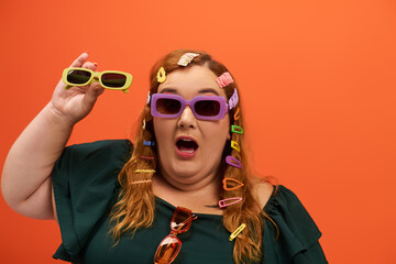 A plus size woman joyfully showcases her colorful style with playful accessories and sunglasses.