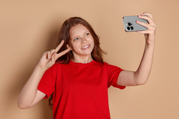 Smiling teenage girl taking selfie and showing peace sign on beige background