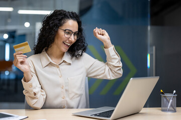 Businesswoman celebrating achievement using laptop in office. Woman holding credit card, smiling, and expressing joy. Concept of success, happiness, and online transaction excitement.