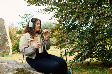 A beautiful plus size woman relaxes in a natural setting while sipping tea from a thermos.