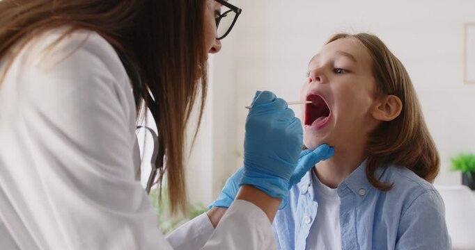 Female smiling pediatrician examining little boy patient's throat in medical clinic during checkup. Doctor woman otolaryngologist exam oral cavity of sick child in office. 4k video. Slow motion video
