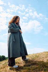 A confident plus size woman enjoys nature while standing in a scenic field on a sunny day.