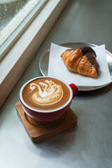 Hot coffee latte with latte art milk foam a swan shape in cup mug with chocolate Croissant on wood desk on top view.As breakfast In a coffee shop at the cafe,during business work concept,Rainy season