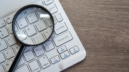 A magnifying glass is placed on a computer keyboard, with the lens hovering over the "Search" key. Online research, investigation, data analysis, or the process of finding information concept.