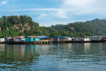 Obraz premium Colorful stilt houses line the riverbank, reflecting a vibrant coastal community. Lush green hills rise in the background under a clear blue sky.