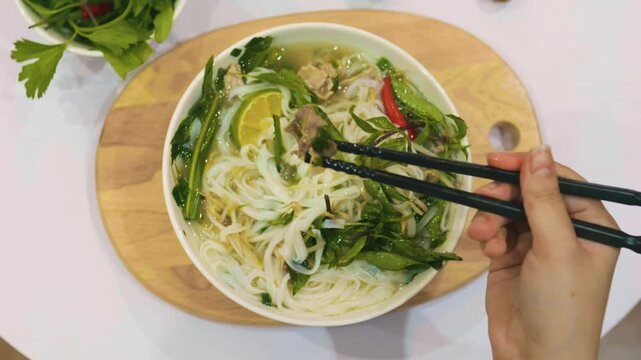 Female hand stirring vietnamese beef noodle soup pho bo with black chopsticks.