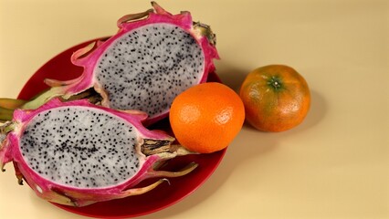 Organic dragon fruit halves reveal speckled white flesh with black seeds beside vibrant orange tangerines on red plate against yellow background. Eco concept emphasizes organic freshness.