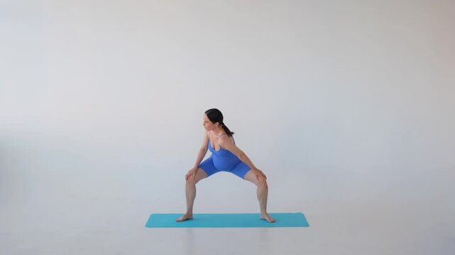 Positive pregnant middle aged Caucasian woman engaged in warm up and stretching shoulder during gymnastic oe pilates training on white background.