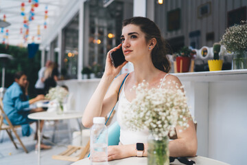 Young woman in activewear sitting at an outdoor cafe, engaged in a phone call. She has a yoga mat and water bottle, surrounded by flowers, enjoying the lively yet peaceful ambiance of the cafe