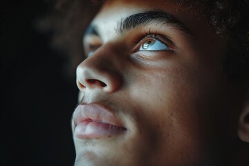 A thoughtful young man gazes upward, deep in contemplation, set against a dark background. The lighting emphasizes his facial features and expression.