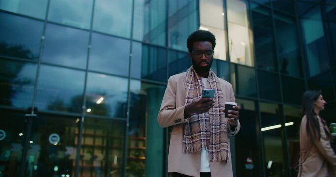 Focused African American male looking at smartphone. Reading news. Swiping on display of mobile phone. Holding cup of hot drink with one hand. Woman passing by in blurred background. Wearing glasses.