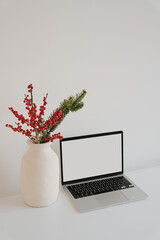 Clean and minimalist workspace featuring laptop with blank screen and holiday decor in white vase with red berries and pine branches. Cozy and modern desk setup