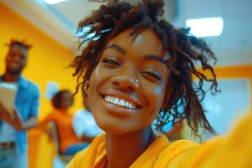 A joyful business team member smiles brightly while taking a selfie in a vibrant office setting with colleagues in the background.