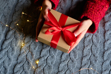 Child's hands holding a Christmas gift box wrapped in brown paper with a red ribbon, set against a cozy gray knitted sweater and surrounded by warm fairy lights.