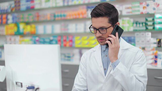 Male pharmacist looking at computer screen. Man druggist working with cash medicine inventory. Man in white gown talking by phone. Showcase with medicine. Caucasian pharmacist in drugstore.