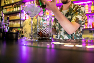 A bartender pours a dark beverage into an ornate glass on a bar counter, with a blurred background of bottles and bar decor. The scene captures the elegance and craftsmanship of cocktail preparation.