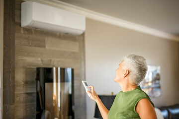 woman holding remote control, adjusting temperature of air conditioner mounted on wall. Indooor...