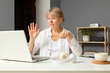 Mature woman with glasses waving hello during video call at her laptop in home office engaging in conversation with colleagues or friends managing her freelance work or conversation online