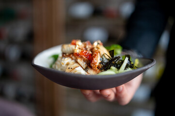 A close-up of a hand holding a bowl of Asian cuisine, featuring rice and vegetables, garnished with sesame seeds and herbs. vibrant colors and fresh ingredients create an appetizing and healthy meal.