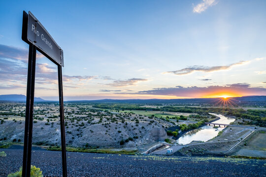 A sign with an arrow pointing to the Rio Grande River overlooking a wide fertile valley as the sun is just setting over a distance mountain ridge, Cochiti Lake Dam, New Mexico