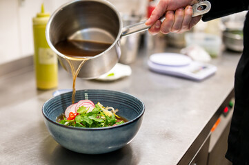 A close-up of a hand pouring clear broth from a metal ladle into a blue bowl filled with noodles,...
