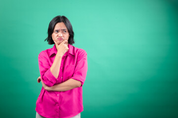 An Asian woman in a bright pink shirt stands against a green background, displaying a pensive...