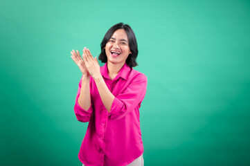 An Asian woman in a bright pink shirt stands against a green background, laughing and clapping her...