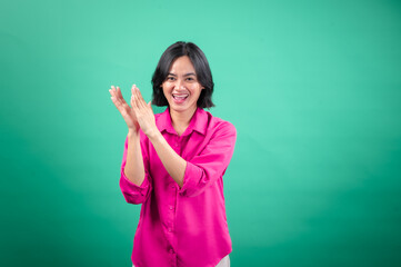 Fototapeta premium An Asian woman in a bright pink shirt stands against a green background, laughing and clapping her hands, showing joy and excitement. Her pose and smile radiate positivity and friendliness.