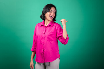 An Asian woman wearing a bright pink shirt stands against a green background, smiling and gesturing...