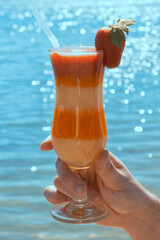 Hand holding a layered tropical fruit cocktail with strawberry by the sea