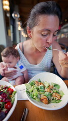 Mother multitasking while eating a salad and holding her baby, balancing caregiving and mealtime at a restaurant during a casual meal out