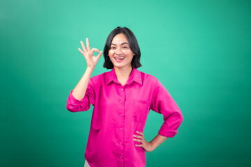 An Asian woman in a bright pink shirt poses against a green background, smiling and winking while making an 