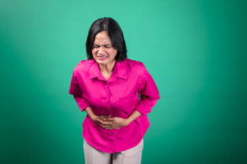 An Asian woman in a bright pink shirt against a green background holds her stomach with both hands,...