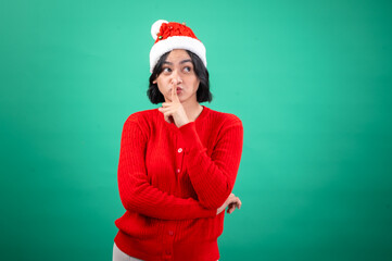 An Asian woman in a red sweater and Santa hat stands against a green background with her finger to her lips, making a 